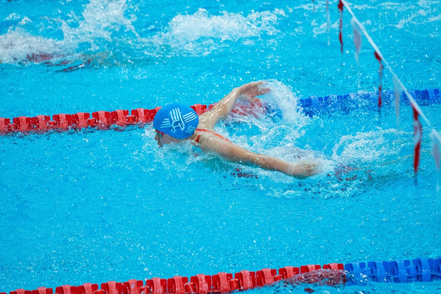 pexels-photo-34676887-34676887 Swimmer in blue cap mid-butterfly stroke in indoor pool with red lane markers.